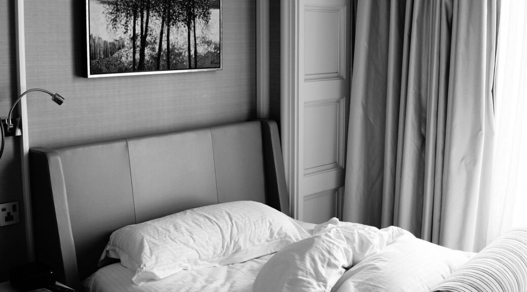 Quiet hotel bedroom with upholstered headboard, curtains, and rumpled white bedding