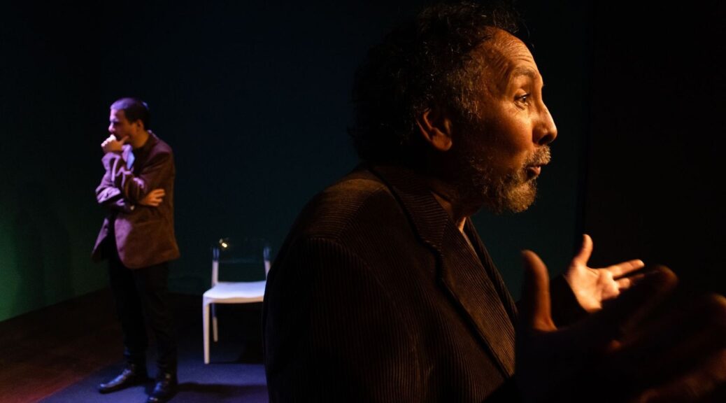 Two actors perform on a dimly lit theatre stage beside a white chair