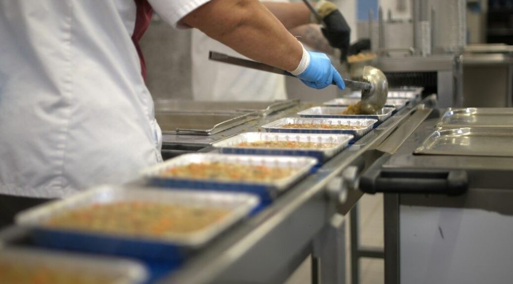 Gloved kitchen worker portions food into trays on a serving line
